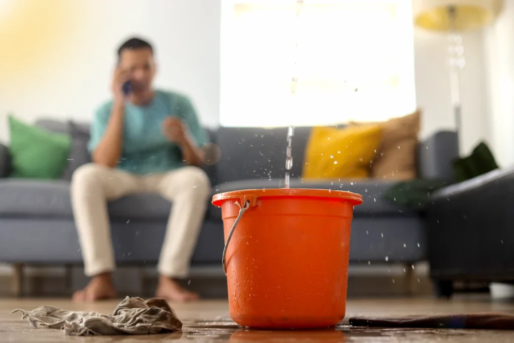Man sitting on a couch talking on the phone while water leaks from the ceiling into an orange bucket placed on the floor, with a wet cloth nearby.