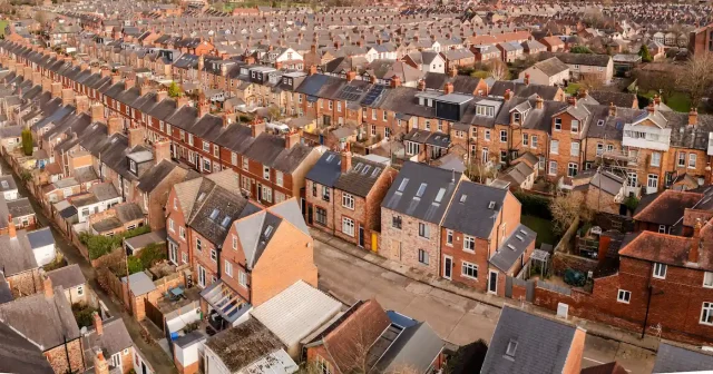 The image shows a dense neighbourhood of traditional terraced houses, typical of many British towns. The houses are made of red brick with slate roofs, and chimneys are visible on almost every rooftop. The streets are narrow and lined with houses closely packed together. Some homes have modern touches, such as skylights or solar panels, suggesting renovations or energy efficiency improvements. The view is aerial, capturing several rows of terraced houses, each with small back gardens or yards. Beyond the immediate row, similar homes stretch into the distance, indicating a tightly-knit residential community.