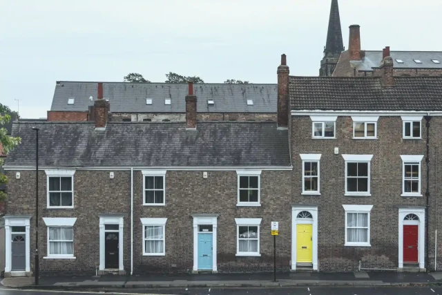 Row of traditional terraced houses with brick exteriors, featuring brightly coloured front doors in grey, blue, yellow, and red. The houses have white-framed windows, and a church spire is visible in the background. The Benefits of Regular Roof Inspections