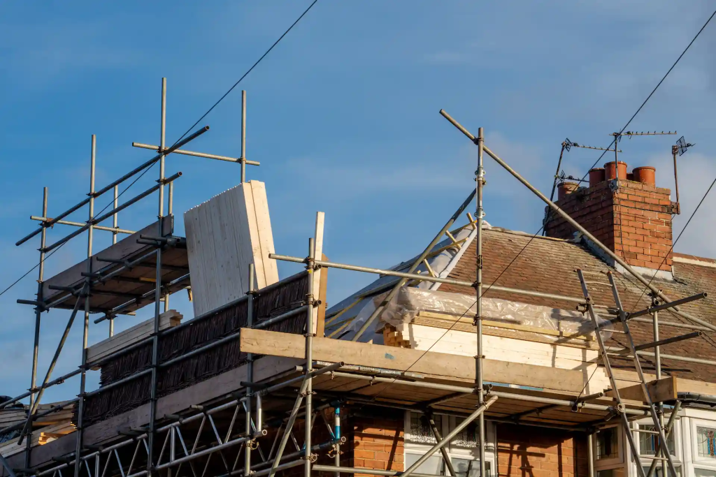A house under construction or renovation with scaffolding surrounding the structure. The roof is partially stripped and covered with wooden panels, plastic sheeting, and insulation materials. Several metal poles and planks form the scaffolding, and a brick chimney is visible alongside a clear blue sky.
