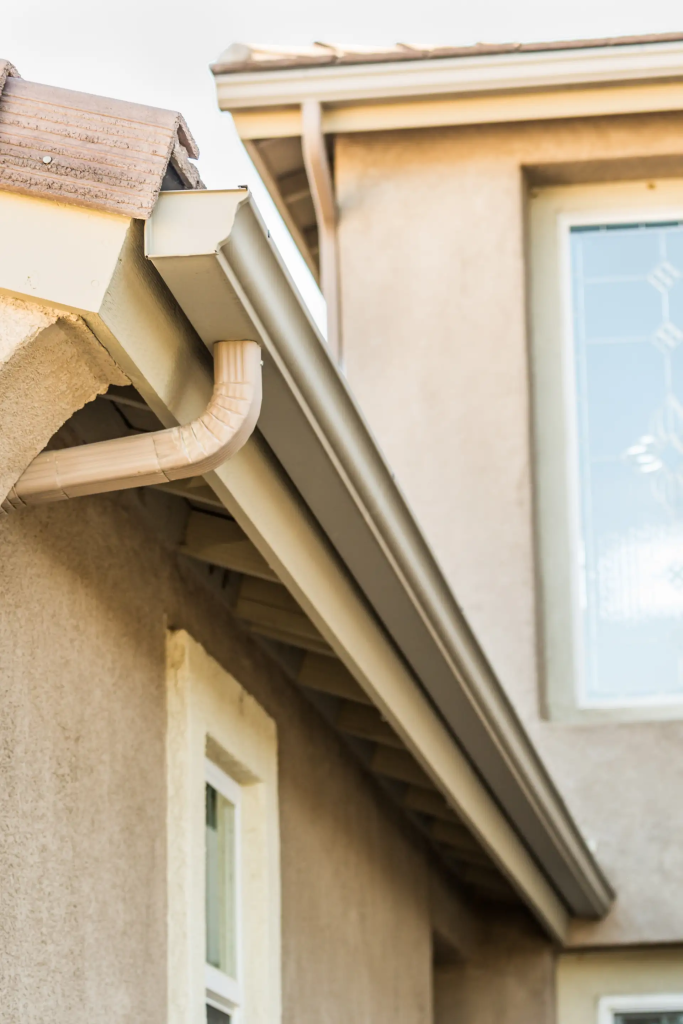 Close-up view of a house’s rain gutter system, including a downspout, attached to the roof of a beige stucco building with a window in the background.