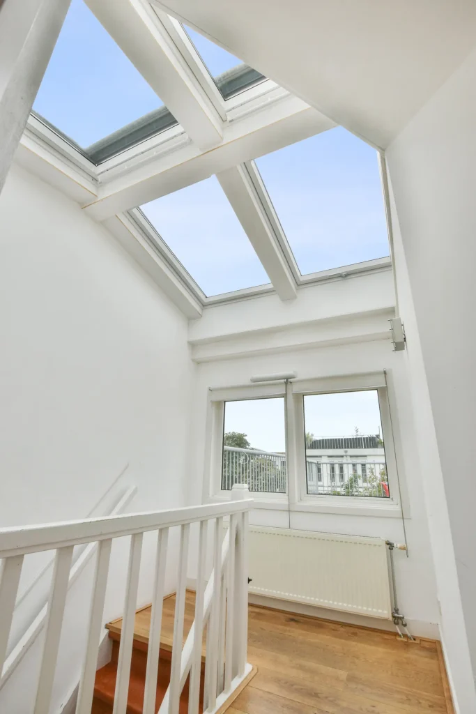 A bright staircase landing with large skylights and windows, allowing natural light to fill the space. The area features white walls, wooden flooring, and a white railing, creating an open and modern atmosphere.